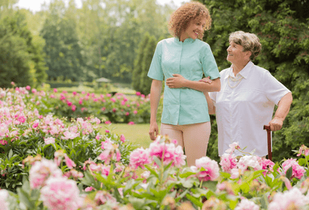 AAA T.L.C. Health Care caregiver walking arm in arm with senior woman among flowers