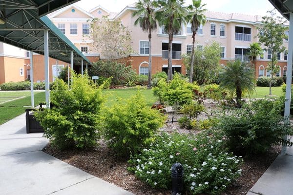 The covered walkway and gardens at Aston Gardens at Pelican Pointe logo