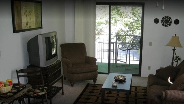 Model apartment living room with a tv, chair, table and a sliding glass door that leads to a patio with a chair.