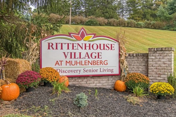 Rittenhouse Village At Muhlenberg sign on brick wall at entrance surrounded by fall colored flowers and pumpkin