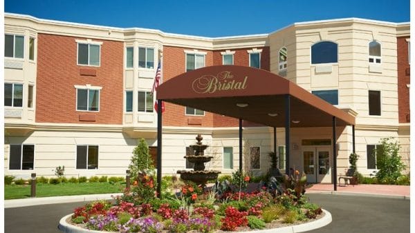 The Bristal Assisted Living at East Northport main entrance with large dark brown awning covering driveway with water fountain and colorful flowers in the rotunda