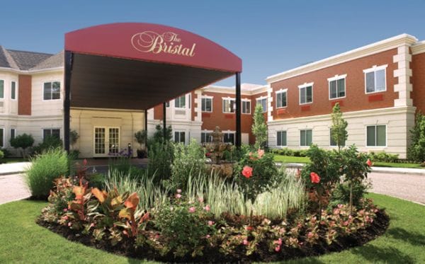 Gardens of flowers in front of the covered awning over the driveway entrance to The Bristal Assisted Living at Massapequa