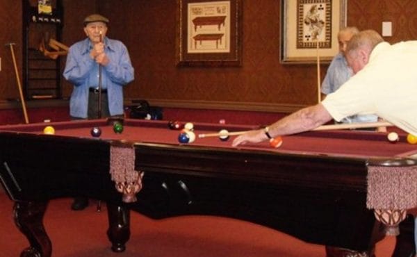 Senior men playing pool on a red billiards table in the gameroom at The Bristal Assisted Living at North Hills