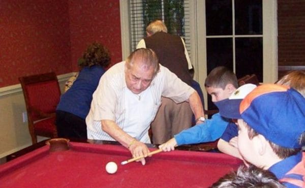 Senior man teaching boys to shoot pool on a red billiards table in the Bristal Assisted Living at North Woodmere gameroom