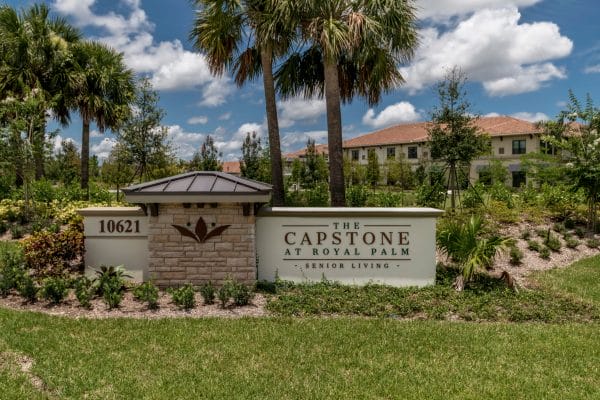 The Capstone at Royal Palm sign at entrance with palm trees and flowers