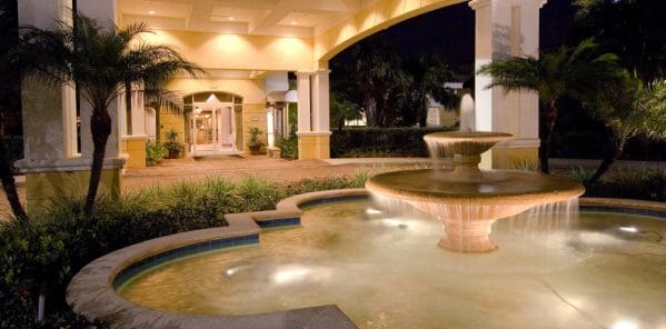 Covered driveway entrance with large water fountain feature at night in front of The Carlisle Naples