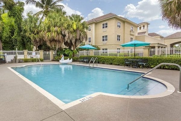 Outdoor swimming pool surrounded by palm trees and umbrella covered tables at Brookdale Bonita Springs