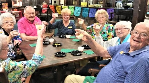 Seven seniors sitting at a table smiling and cheering with a drink in hand