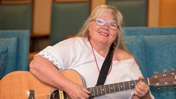 Woman with blonde hair and glasses playing guitar entertaining residents of Parkside Village Retirement