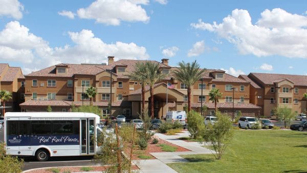 Red Rock Pointe Retirement exterior view with grand palms, community bus and walkways leading up to covered entrance