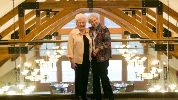 Two senior women standing on balcony at Reflection Ridge Retirement with exposed rafters of vaulted ceiling over lobby behind them