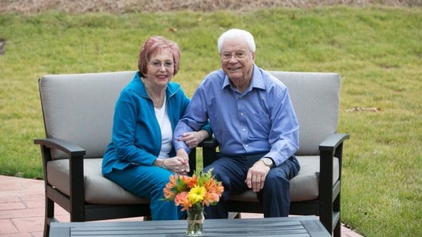 Senior couple holding hands and smiling seated on cushioned chairs outside of Sherrill Hills Retirement