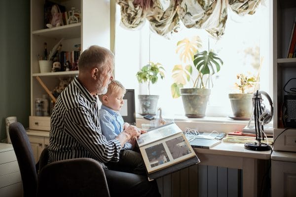 Senior male parent looking at photo memories album with future generation male toddler in front of a window