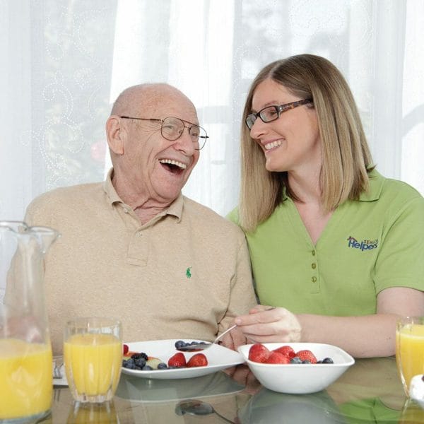 Senior man at a dining table being assisted by a Senior Helpers caregiver and laughing