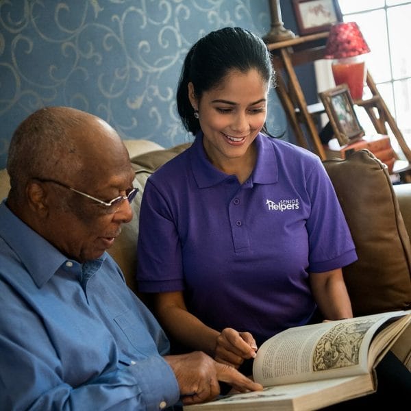 Female Senior Helpers caregiver reading through a book with a senior man on the couch