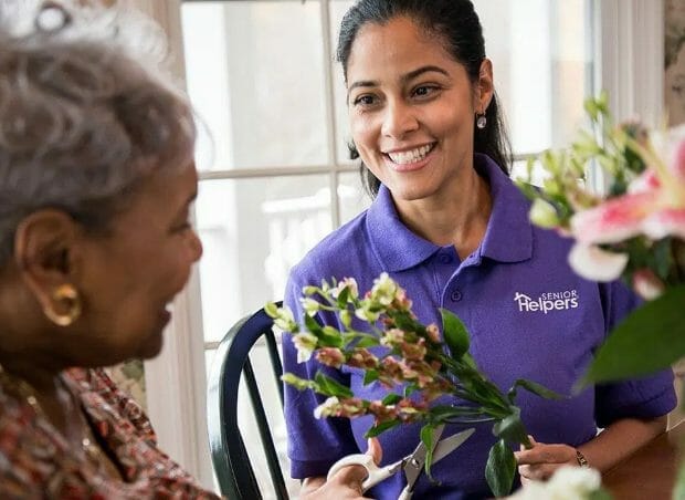 Female Senior Helpers caregiver in purple shirt arranging flowers with senior woman