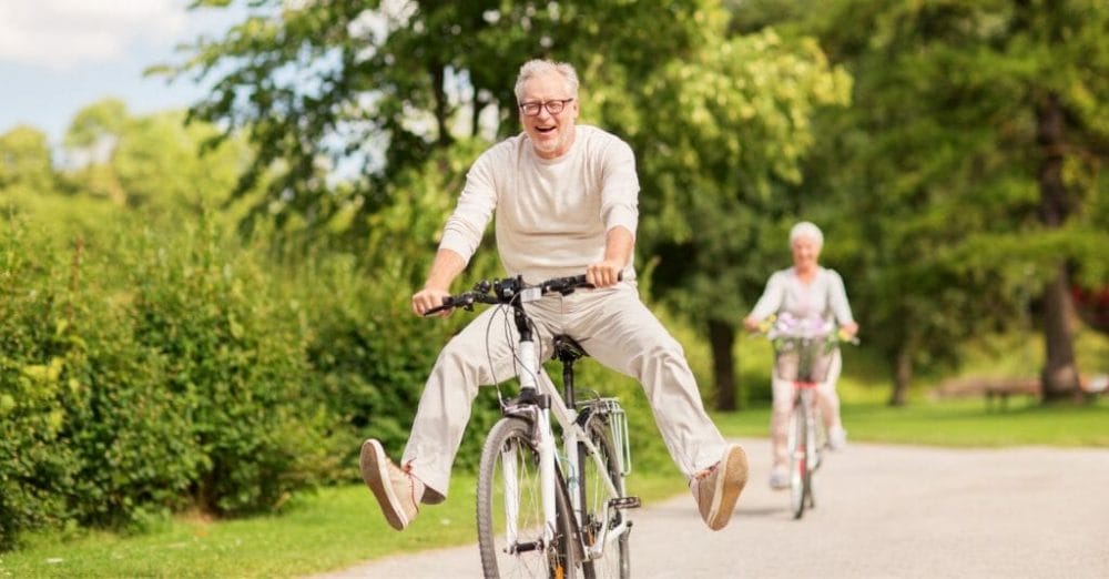 Senior man on a bicycle with his feet off the pedals and laugning while a senior woman rides a bike behind him