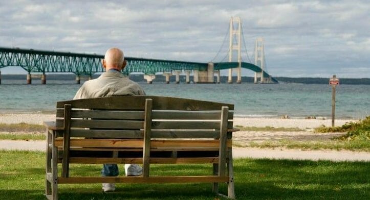 Senior man on park bench in Michigan looking at a long bridge over water
